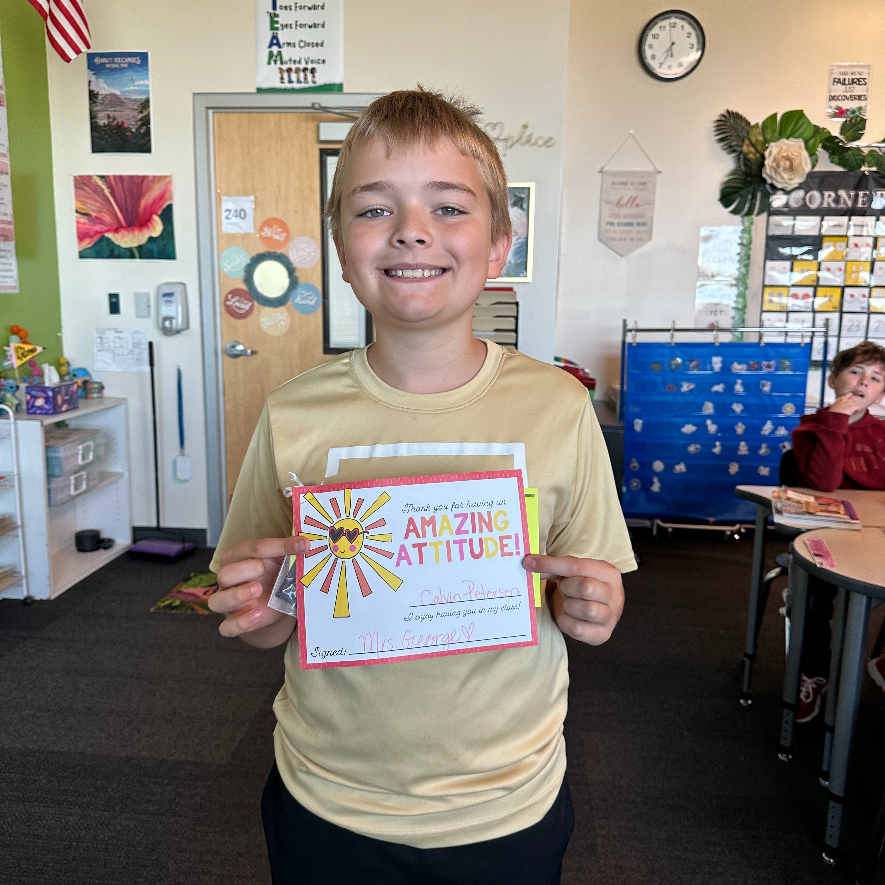 Boy holding a certificate stands in a classroom. Behind him is a door and a clock on the wall.