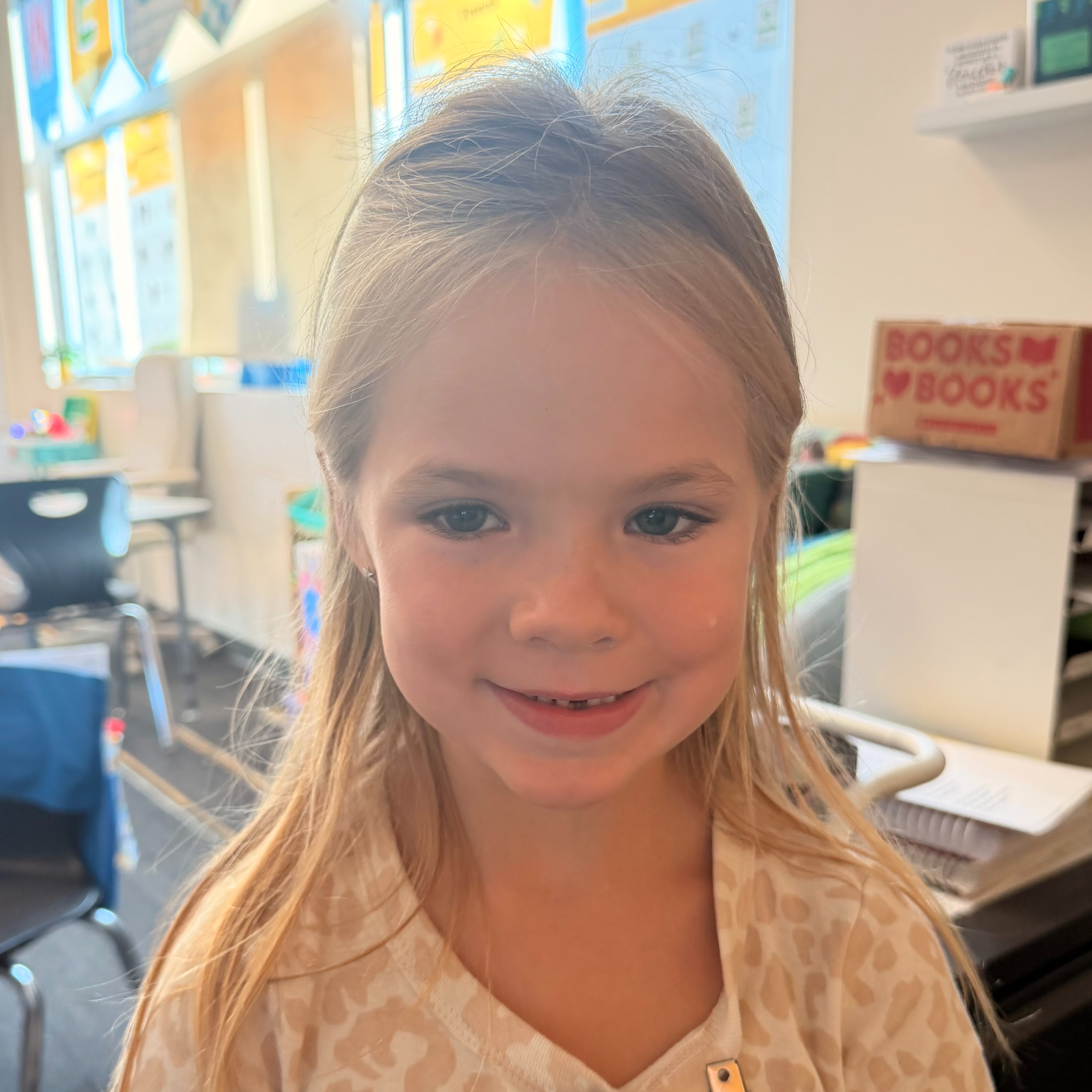 A girl with blonde hair, wearing a leopard print shirt, smiles at the camera in a classroom.