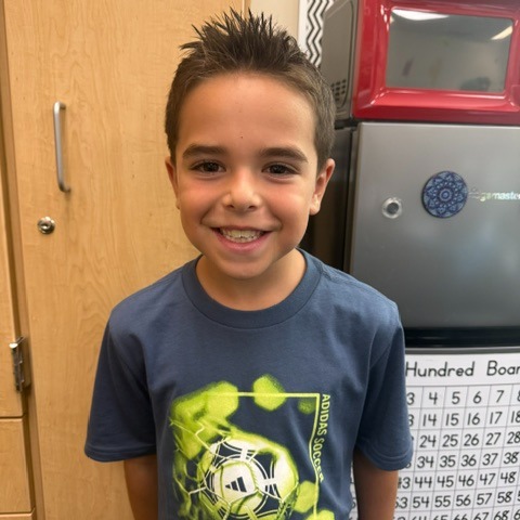 A young boy with spiky hair smiles in front of a classroom door, wearing a blue t-shirt with a soccer design.