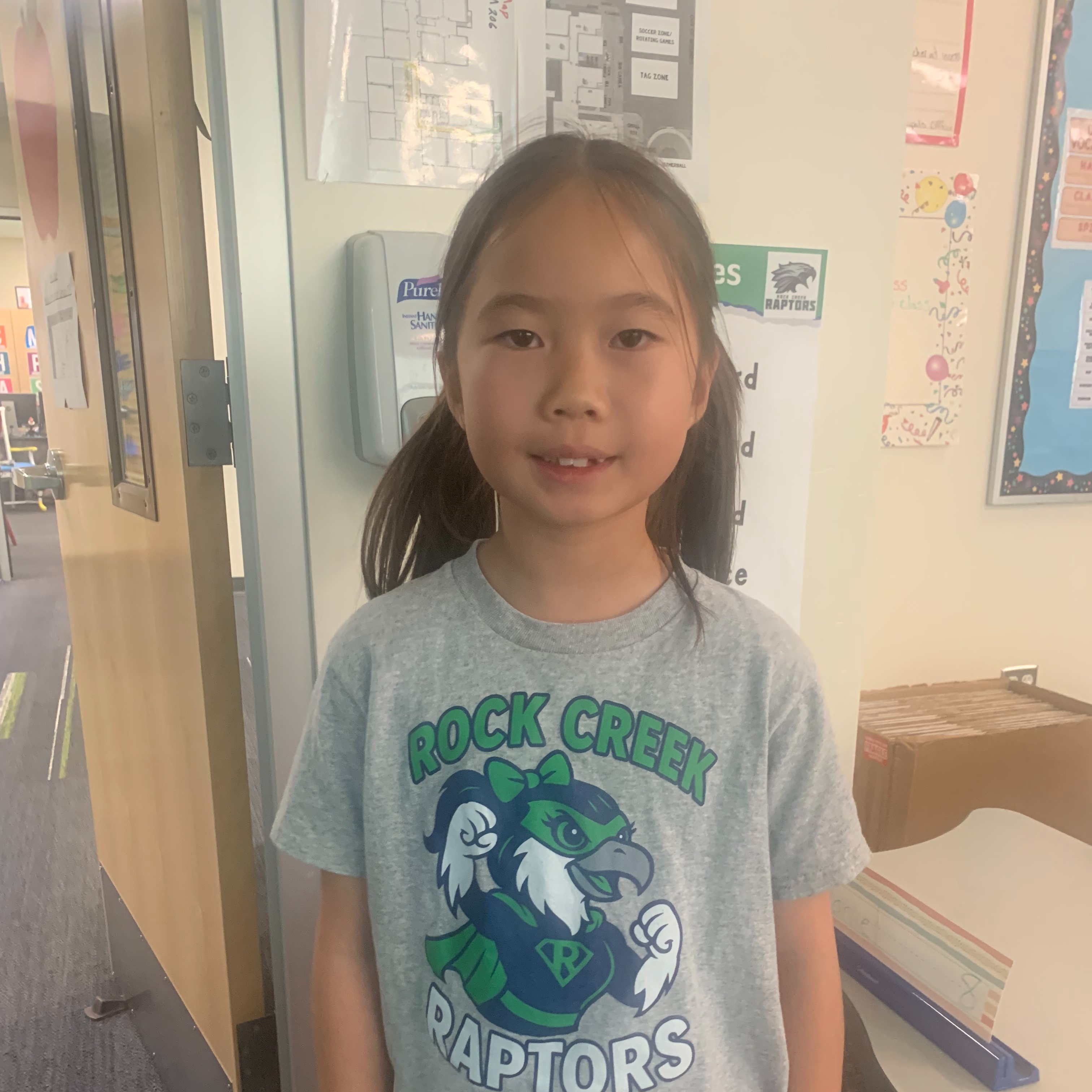 A young girl in a gray t-shirt with a green graphic and pink pants stands in a classroom.