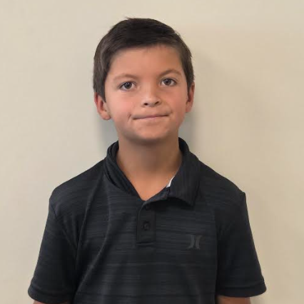 A boy in a black polo shirt stands against a white background, smiling with hands by his sides.