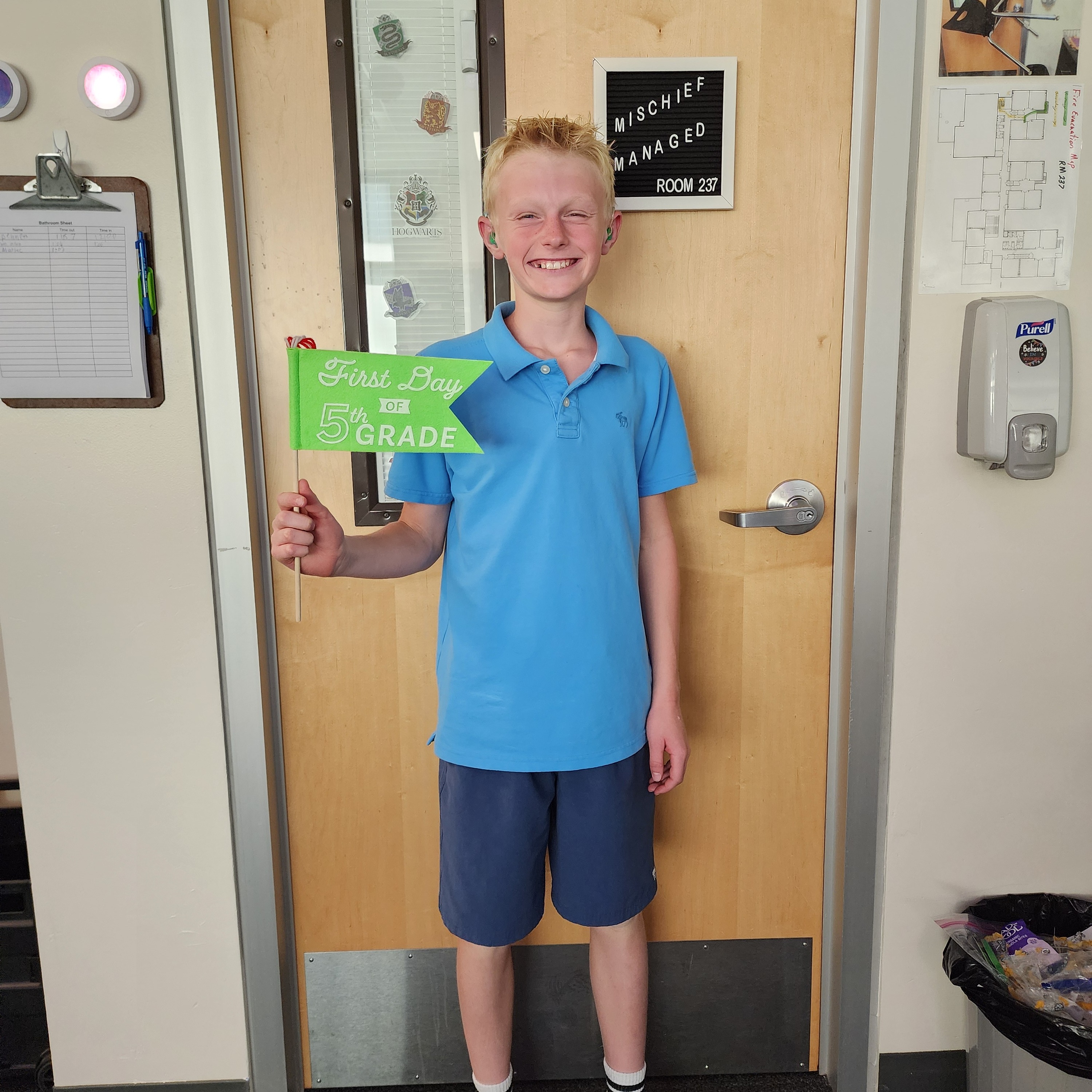 A boy in blue shorts and sandals holds a green sign reading "GRADUATE". He stands in front of a door.