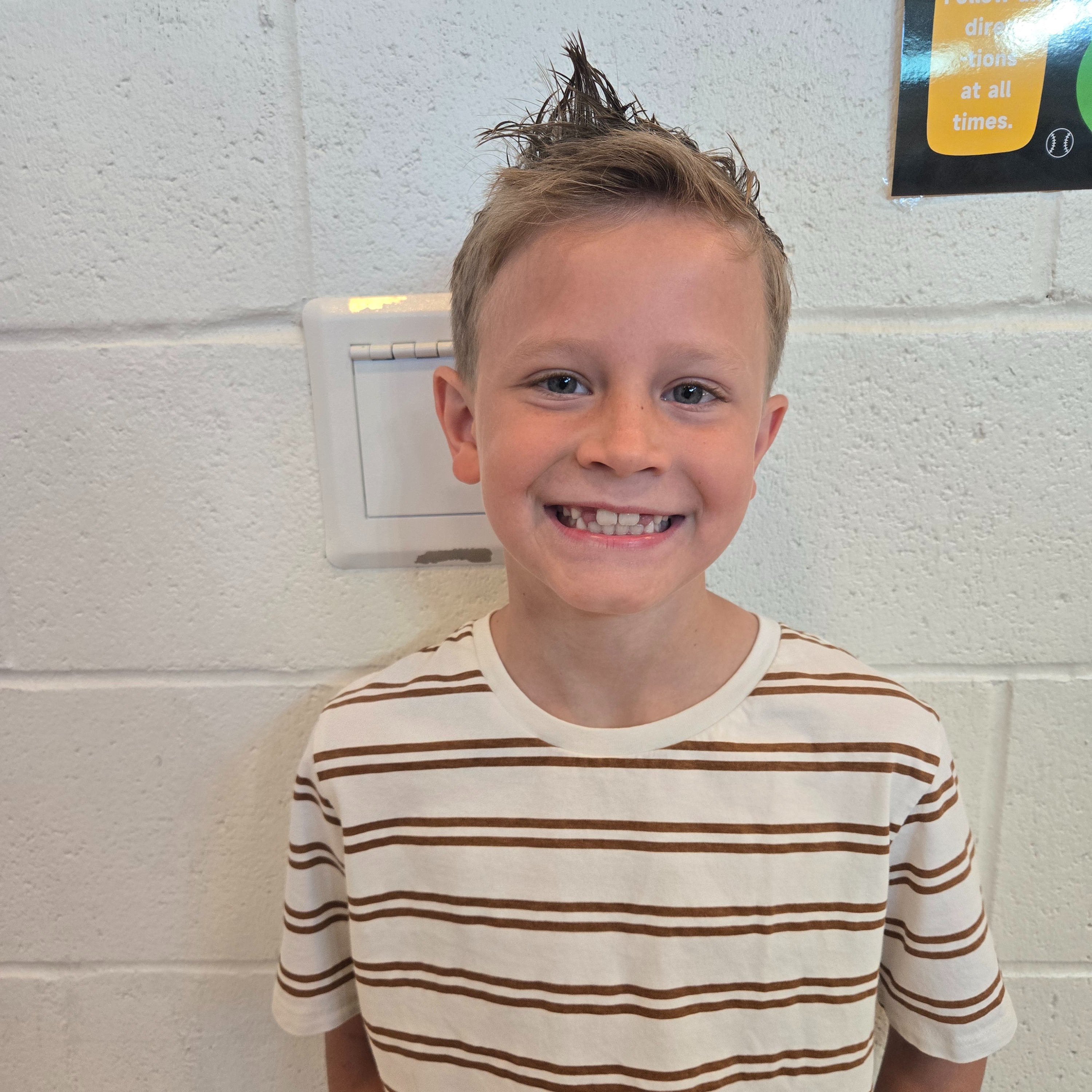 A boy with tousled hair, smiling, stands in front of a white wall. He wears a striped shirt.