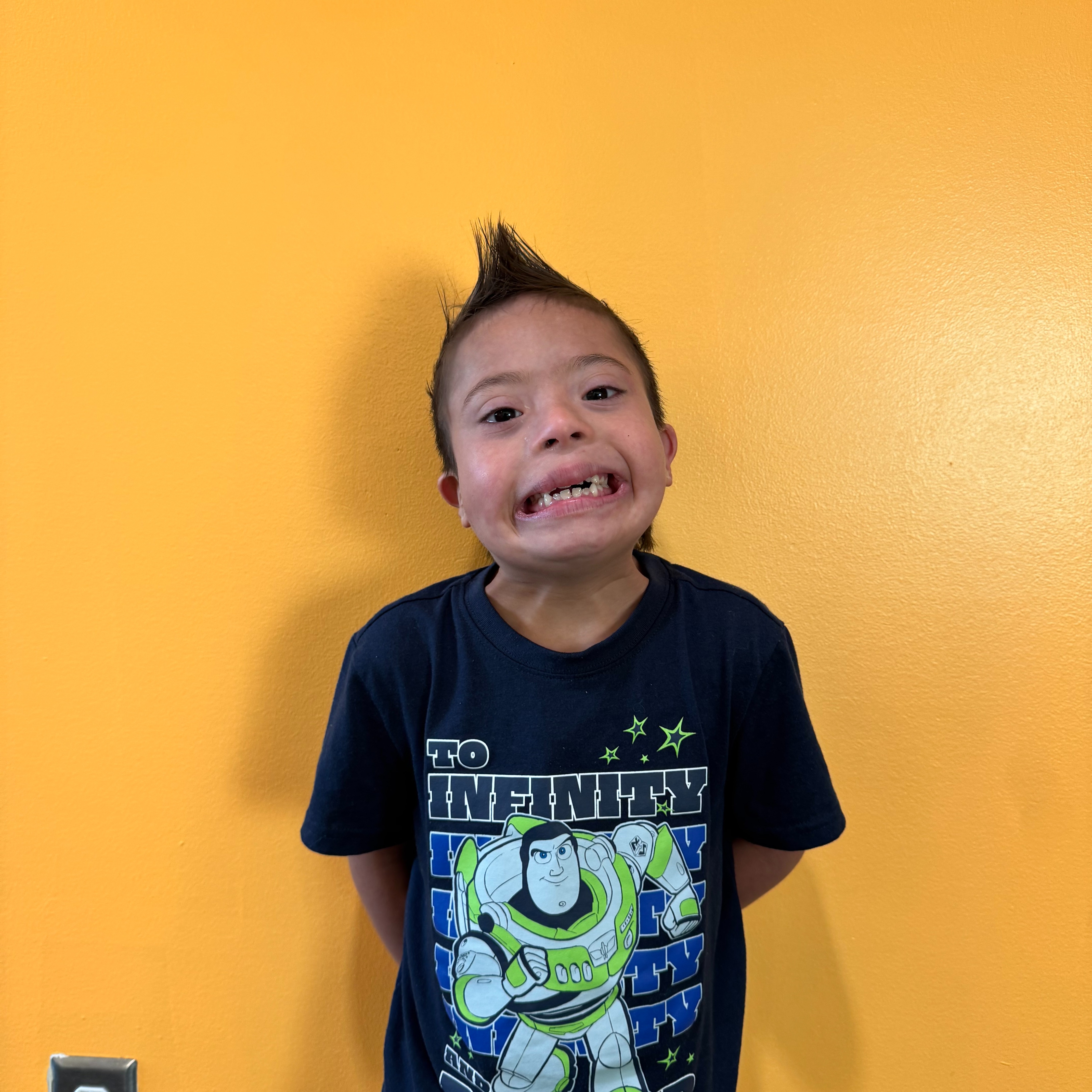 A smiling young boy with spiky hair stands against a bright yellow wall wearing a Toy Story t-shirt.