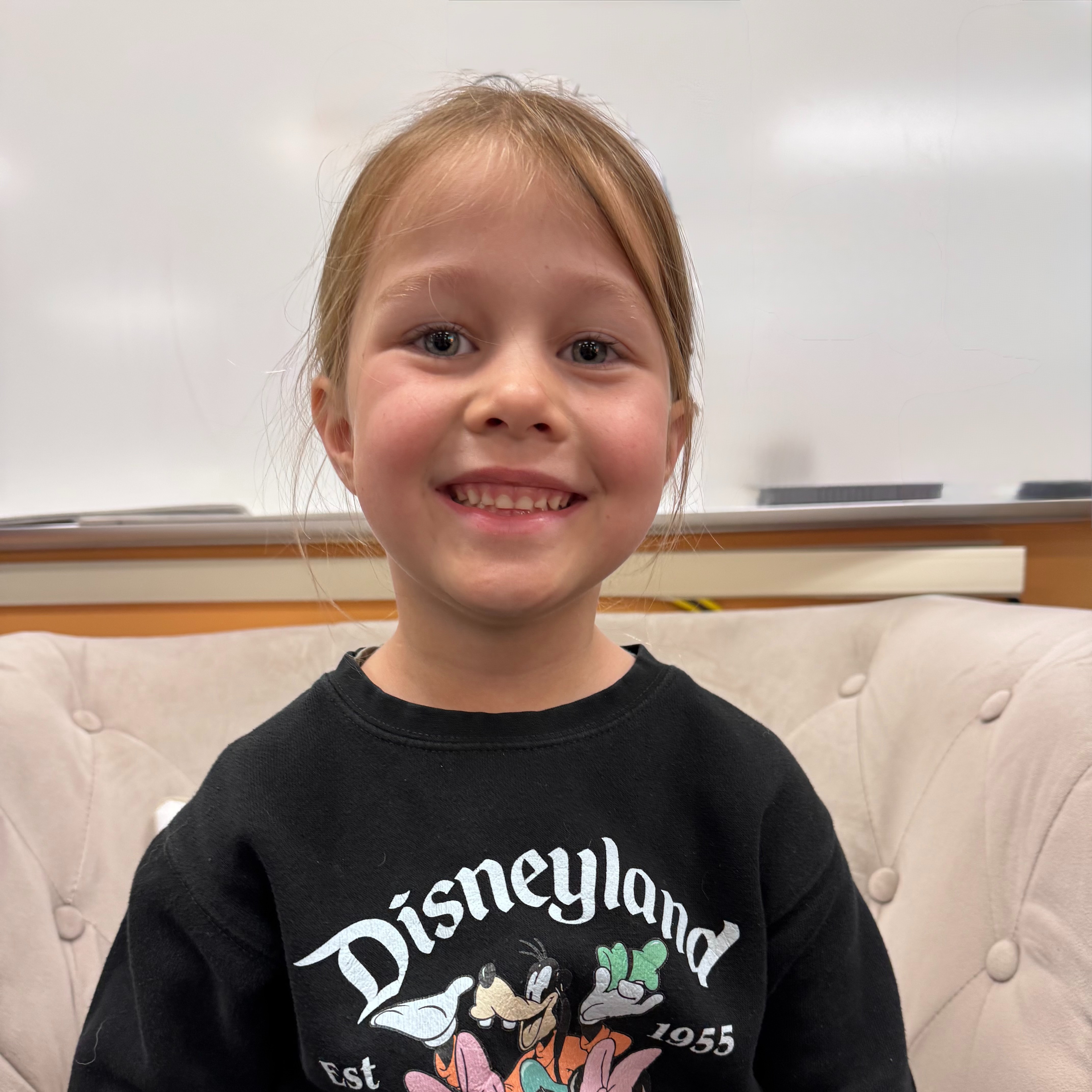 A child with blond hair wearing a black Disneyland sweatshirt sits on a couch. The background features a whiteboard.