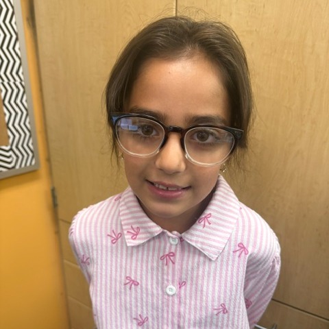 A young girl in glasses stands, smiling, in front of wooden cabinets. She wears a pink shirt with white stripes.