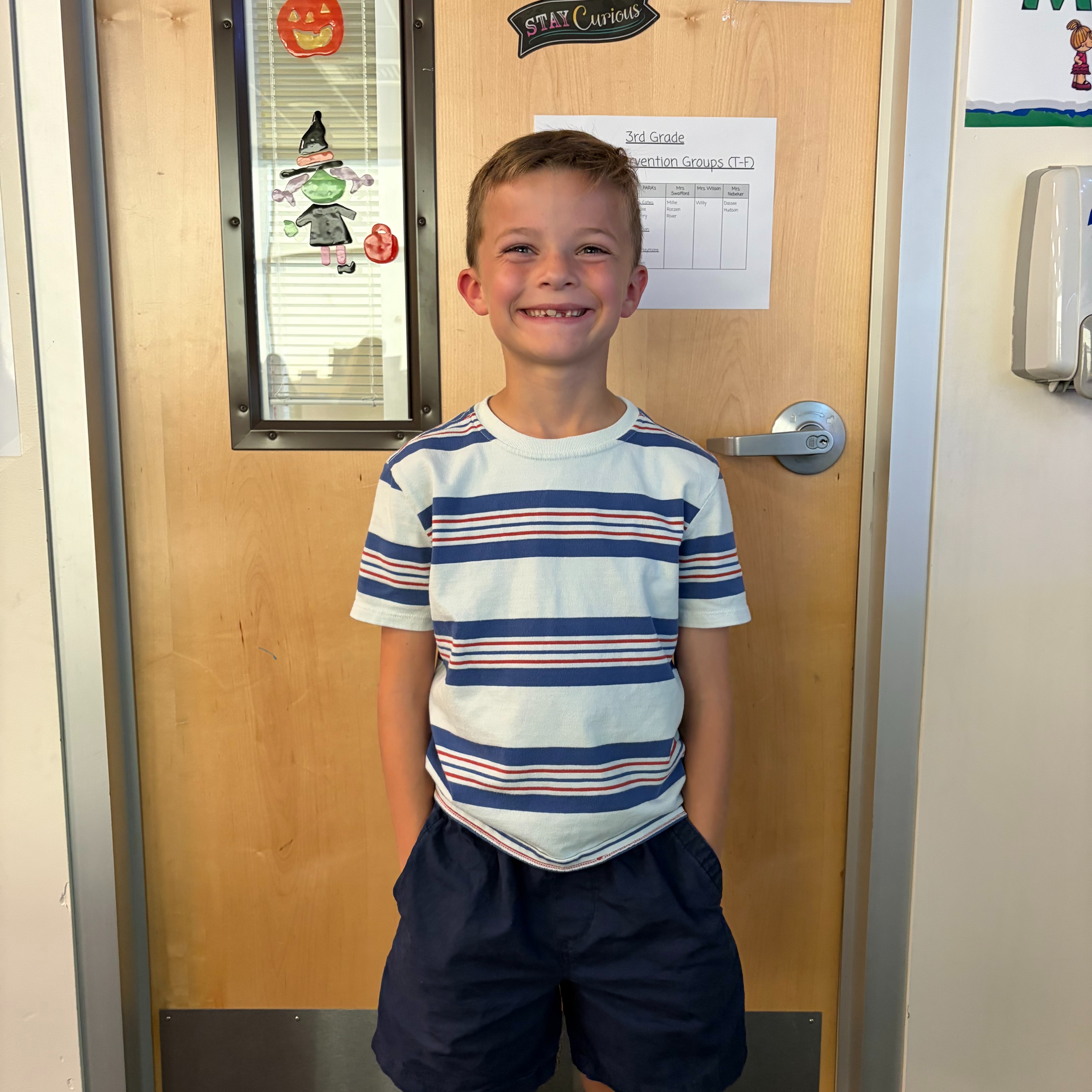 A child stands by a door with "205" on it. They wear a striped shirt and shorts. Behind them is a glass window.
