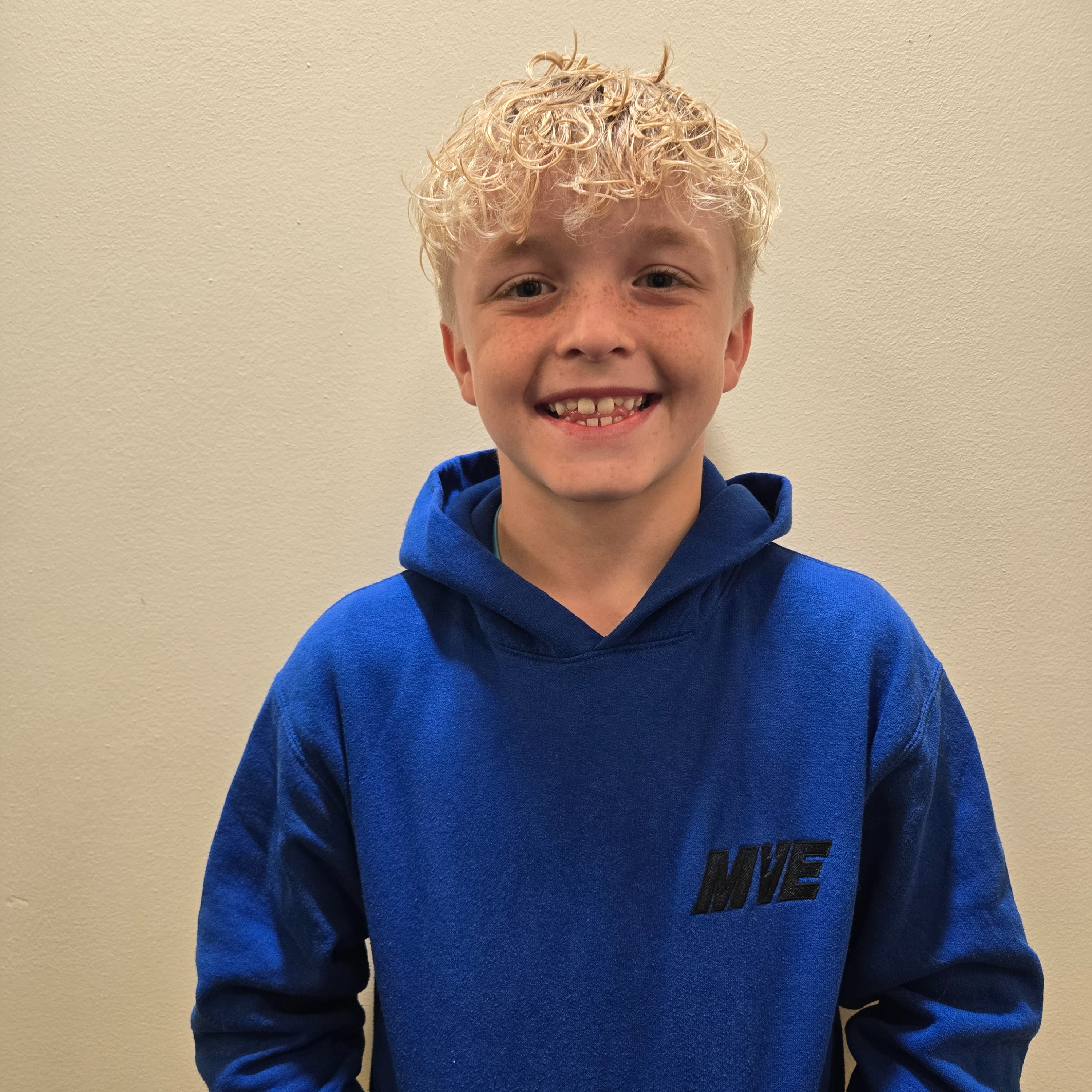 A child with blonde hair smiles at the camera, wearing a blue hoodie with black lettering on the front.