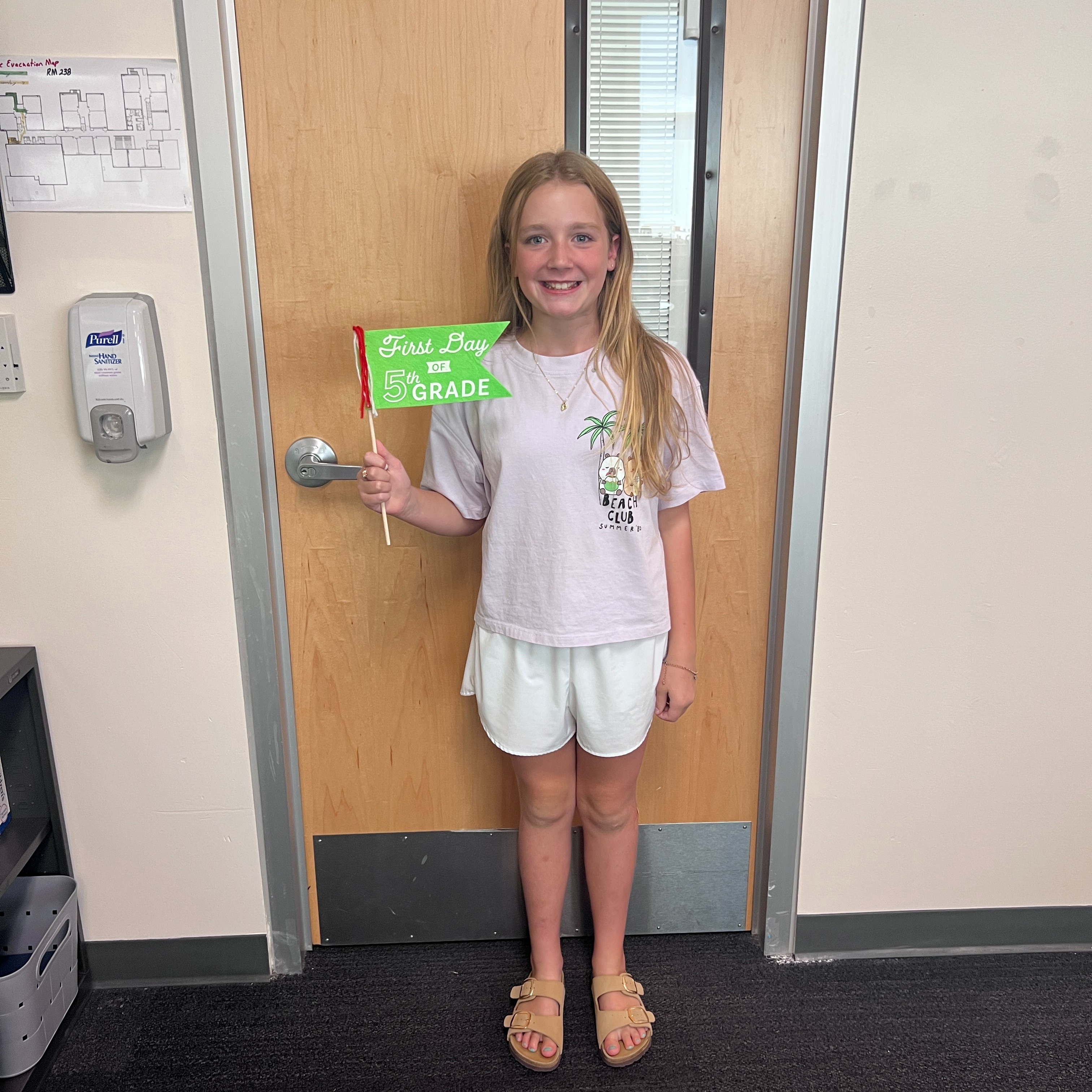 A young girl stands in a doorway holding a green banner with white text. She wears a pink shirt, white shorts, and sandals.