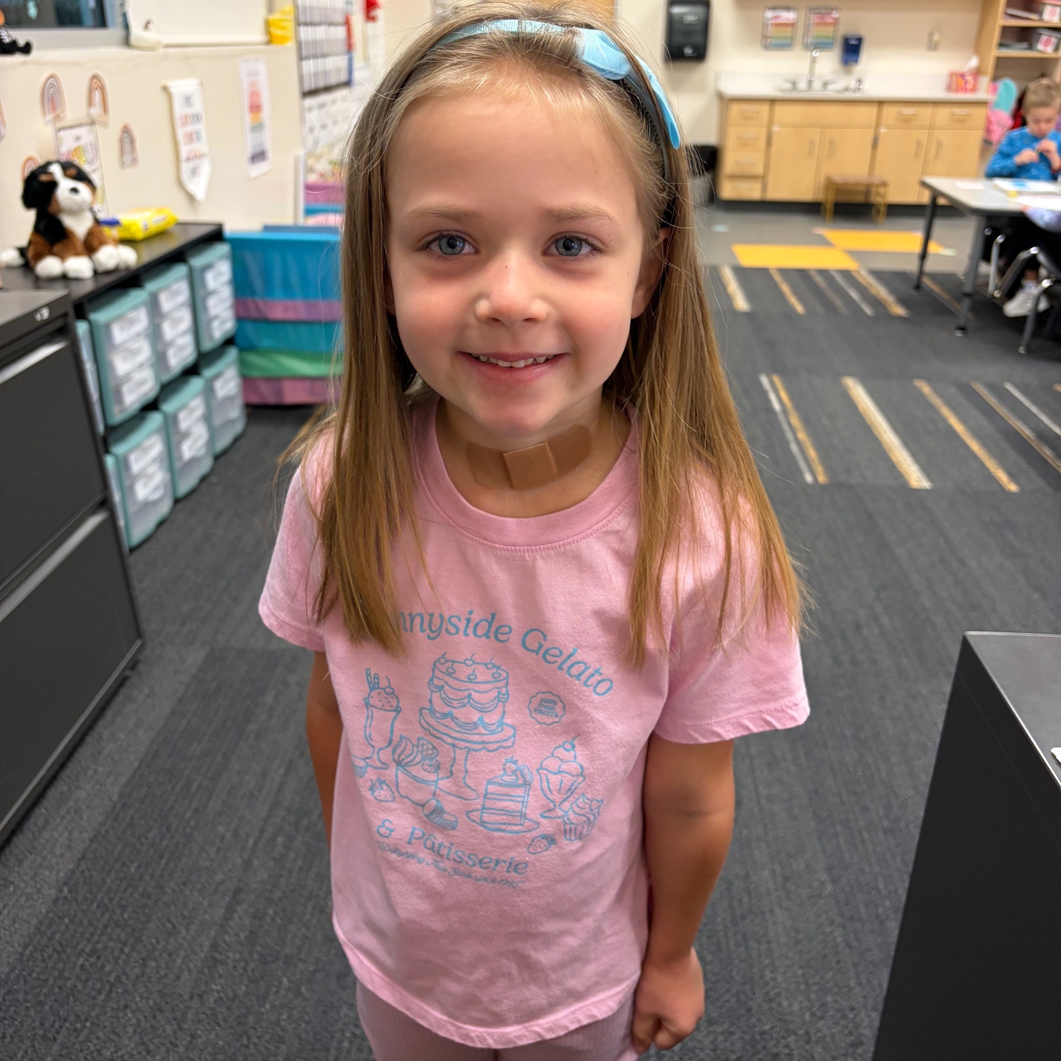 A young girl with long hair smiles for a picture in a classroom. She wears a pink shirt and pink pants.
