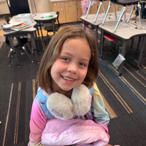 A young girl sits in a classroom with a blanket around her shoulders, smiling.