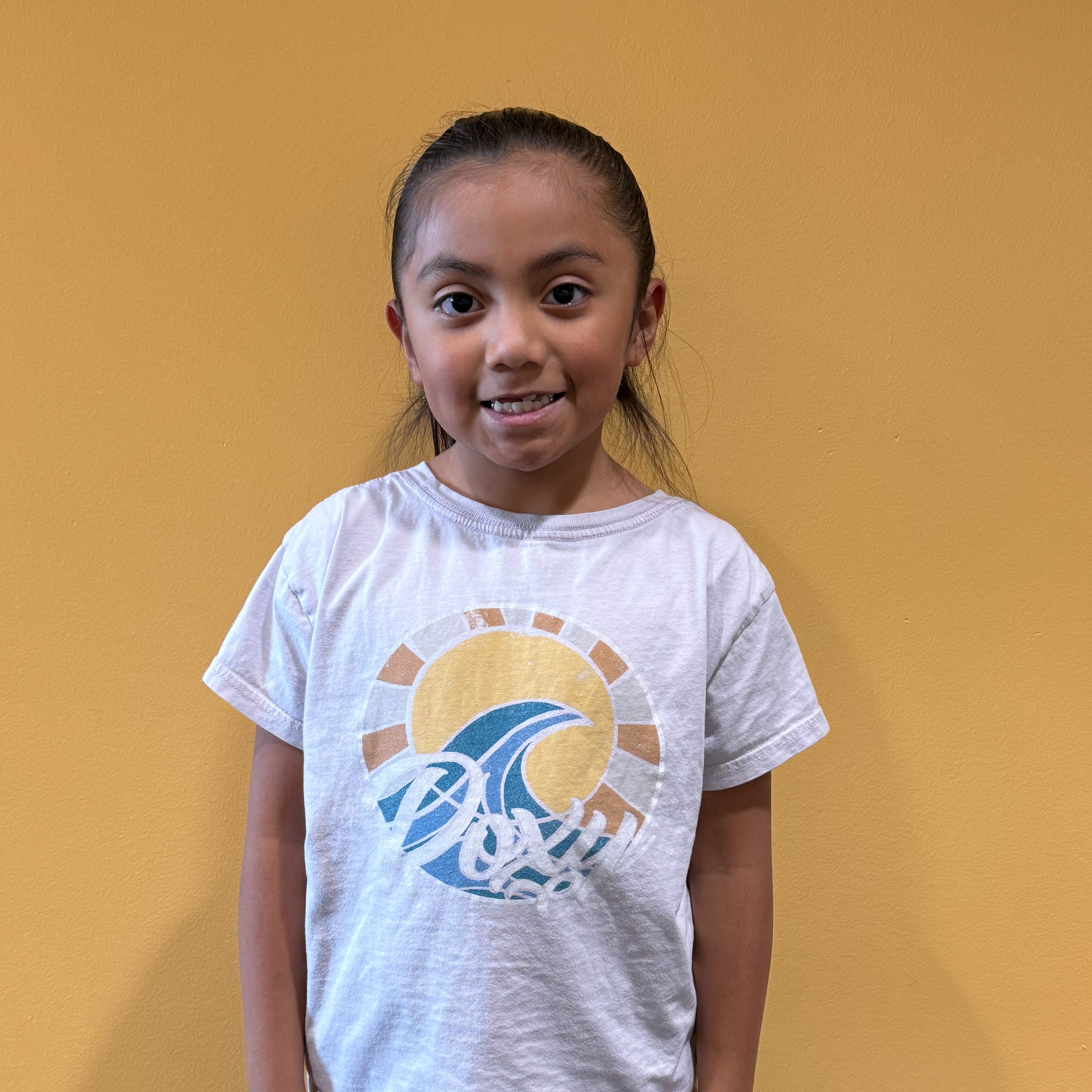 A girl with short hair smiles while standing in front of a yellow wall. She wears a white t-shirt with a blue design.