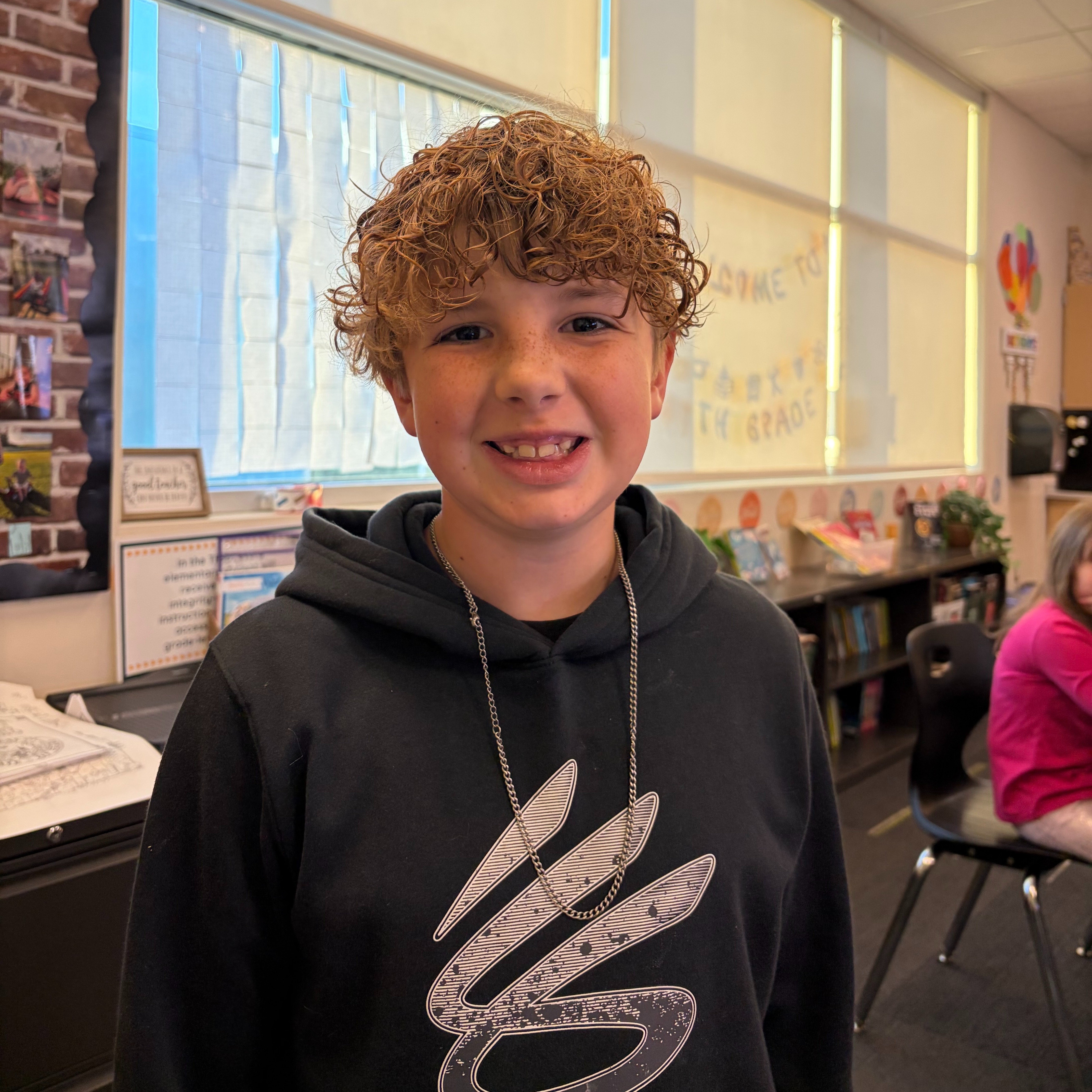 Brightly lit classroom with windows. Young boy with curly hair stands and smiles. Behind him, a person sits at a desk.