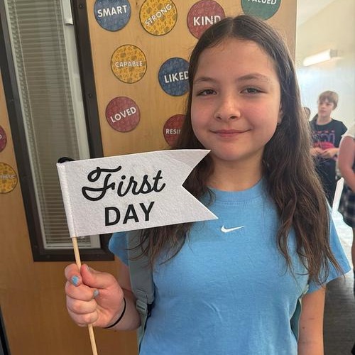 Young girl in light blue shirt holding a white banner saying "First Day" in front of a bulletin board.