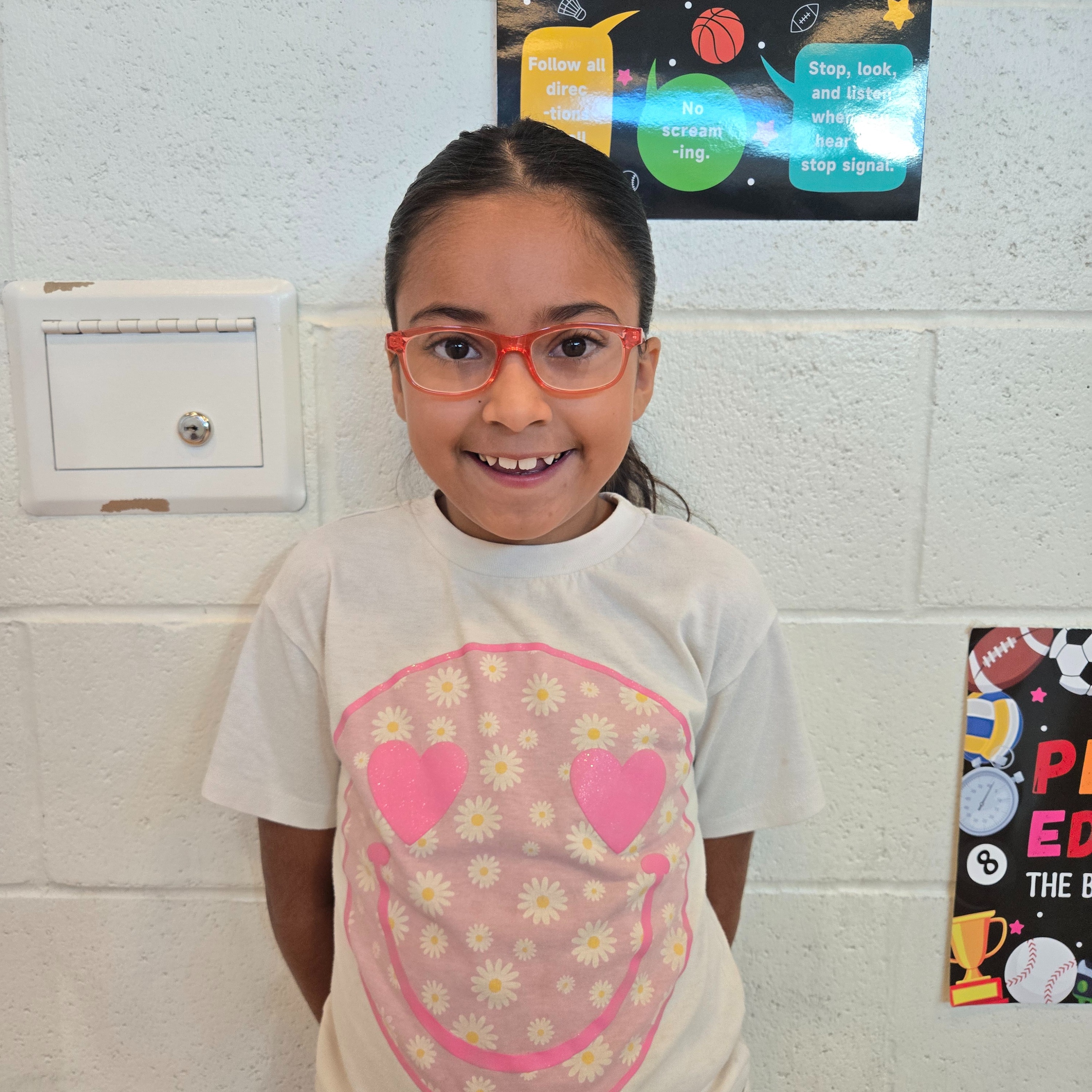 A smiling child wearing glasses stands in front of a white wall. There are posters in the background.