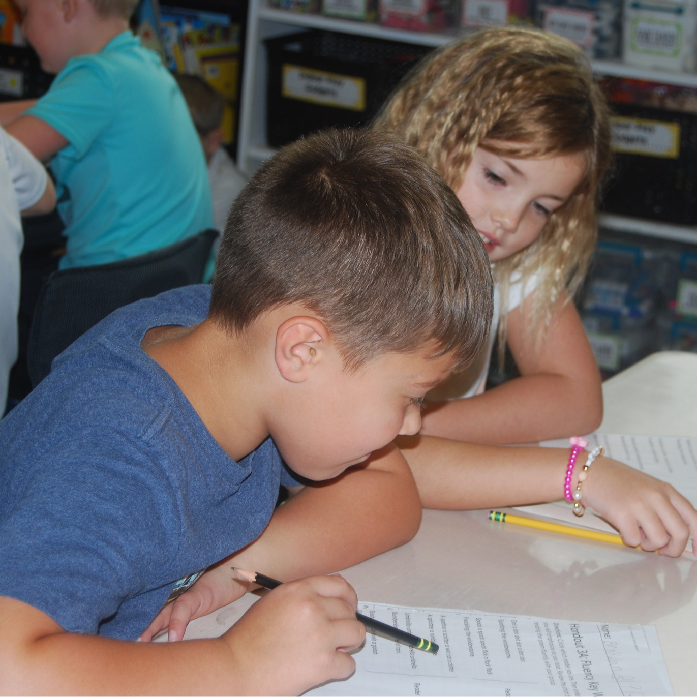 Photo of an elementary aged boy in blue shirt holding pencil looking at a worksheet. There is an elementary-aged girl sitting behind him looking over his shoulder at the worksheet.