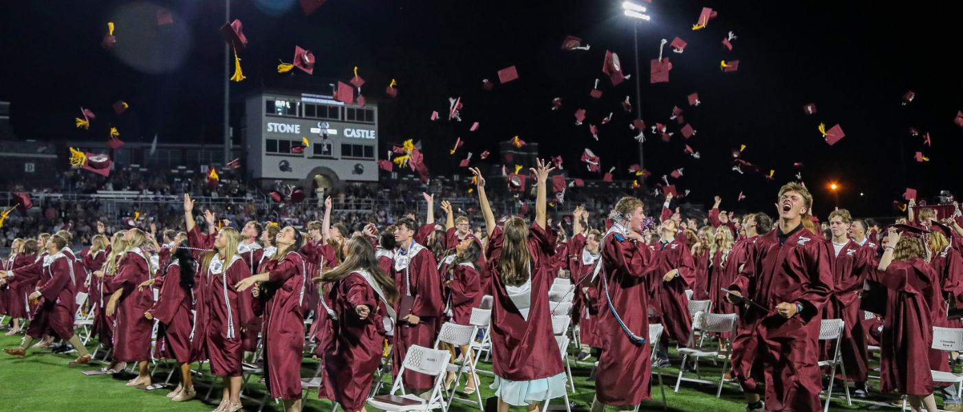 Tennessee High School graduates in maroon caps and gowns celebrate on the football field at night by tossing their caps into the air after commencement. Stadium lights shine over the crowd as students cheer, raise their hands, and celebrate together in front of the “Stone Castle” press box and packed bleachers.