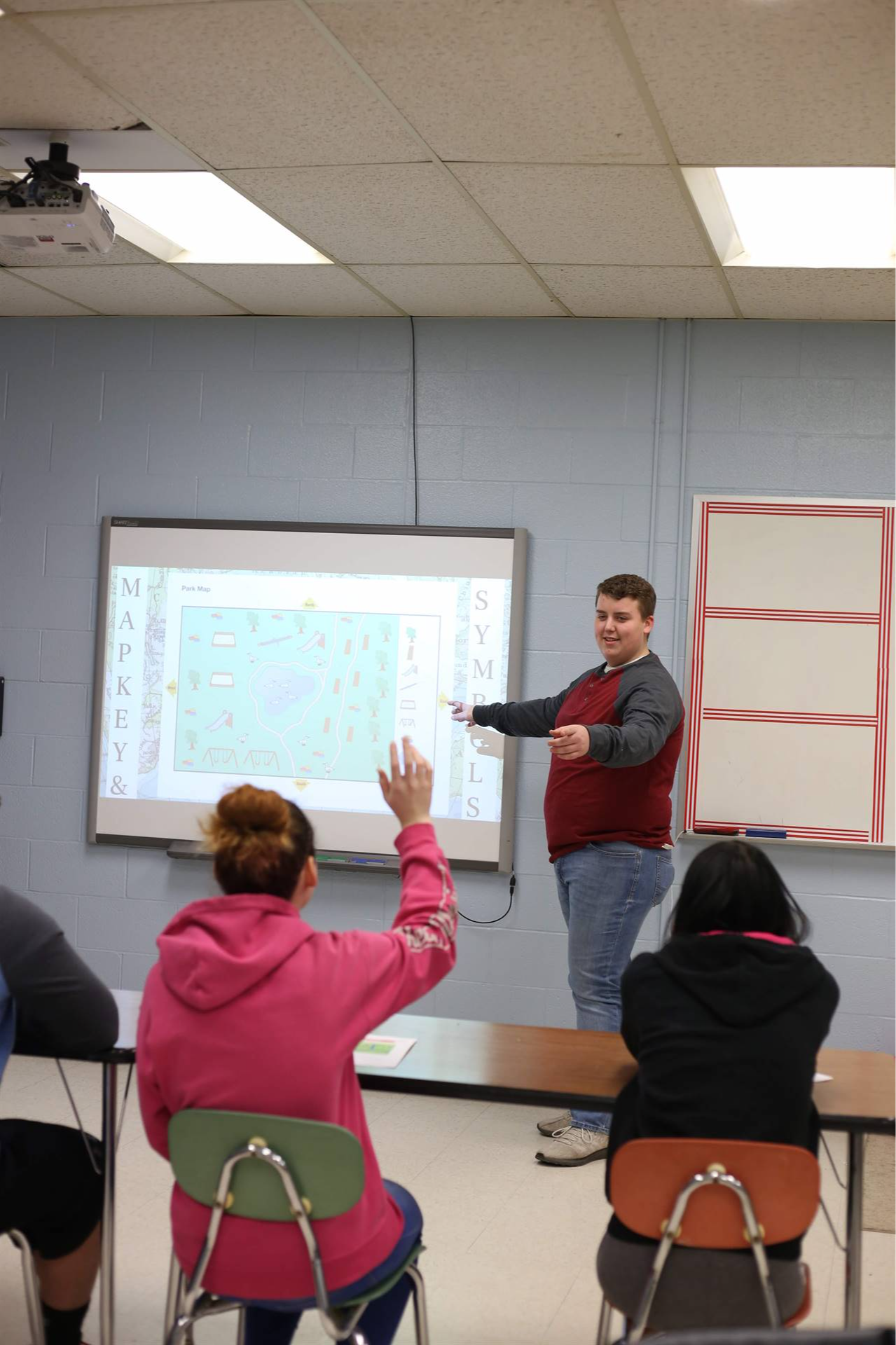 student at the front of the classroom pointing at a girl with her hand raised