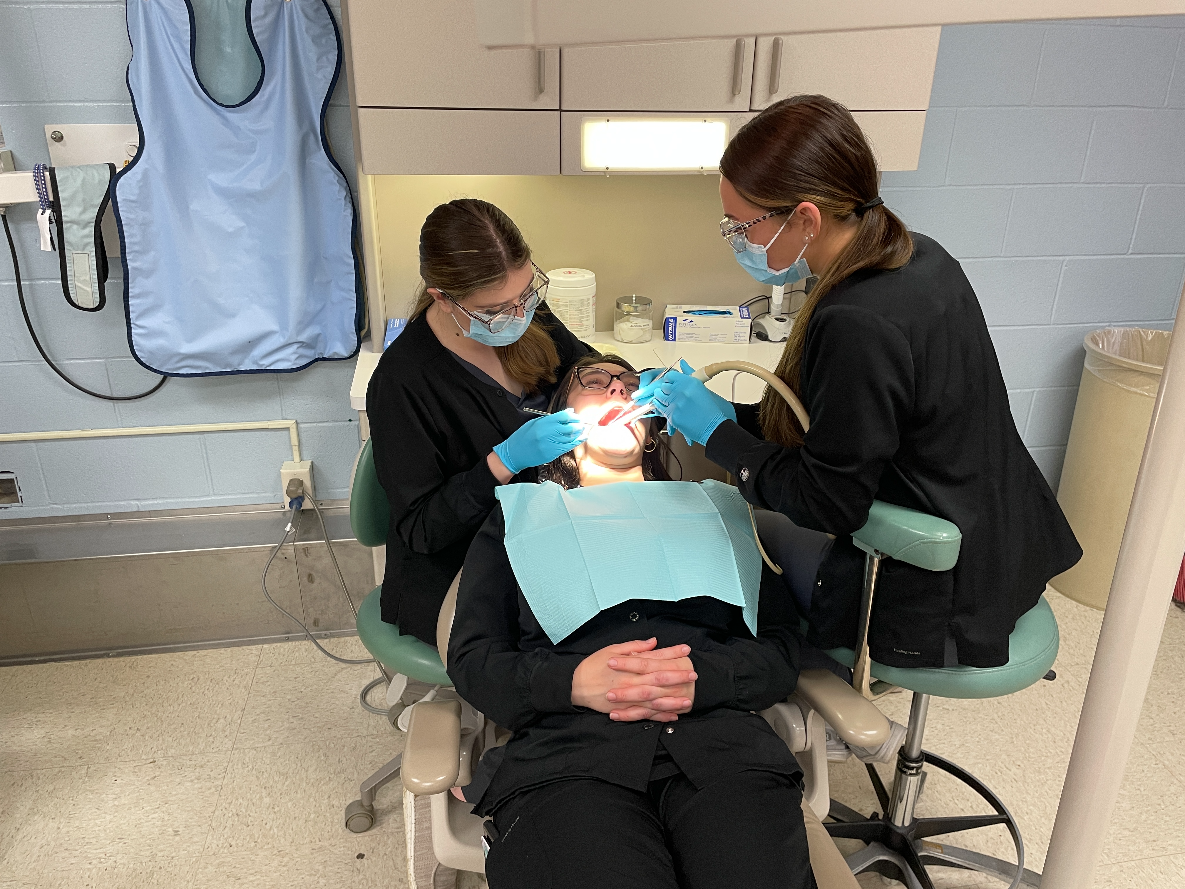 2 adult students looking in mouth of another student as she sits in dental chair