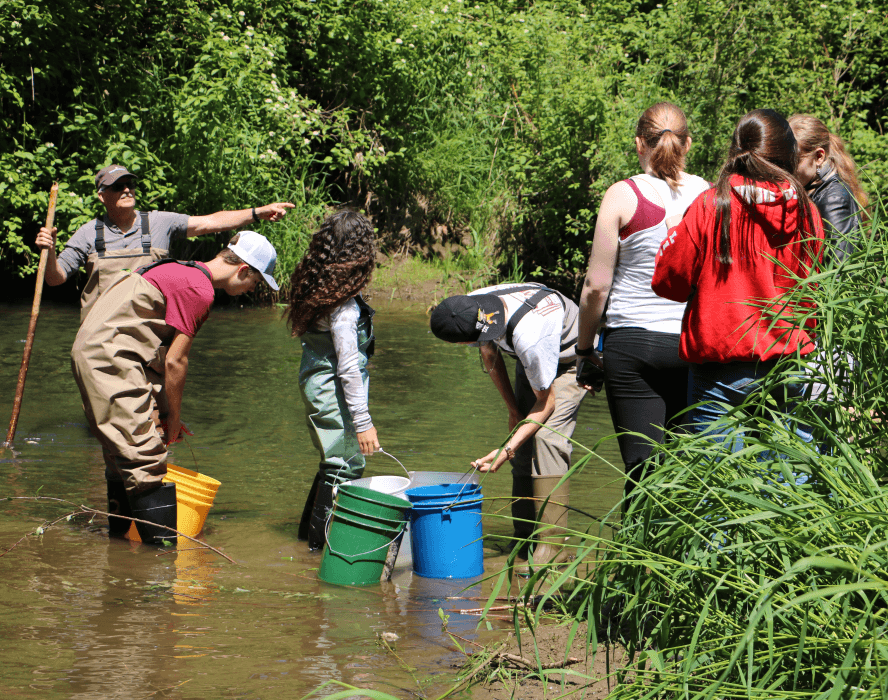 A group of people standing near a body of water, with some fishing and others observing.