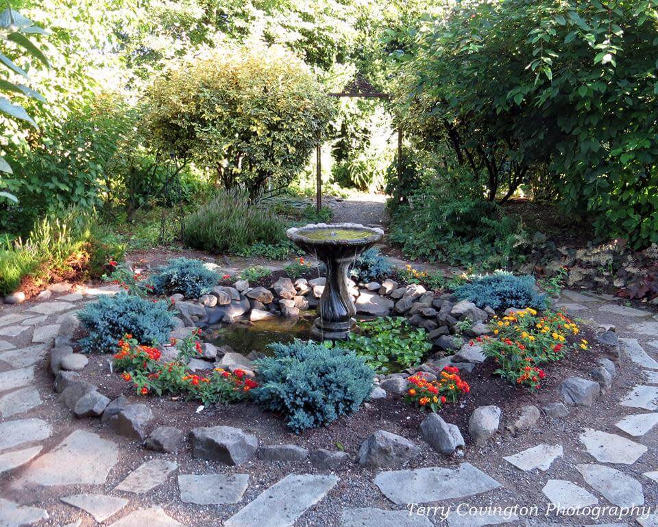 A tranquil garden oasis featuring a central fountain surrounded by colorful flowers and bordered by a peaceful pond.