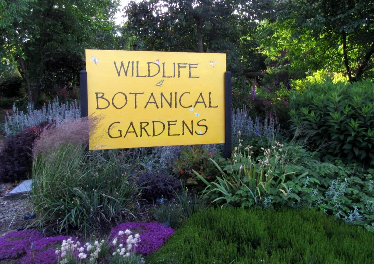 A yellow sign in a garden, reading 'Wildlife Botanical Gardens'.