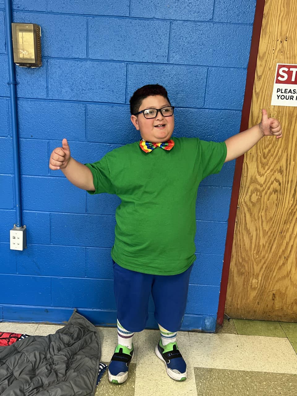 A boy in glasses and a green shirt smiling and giving a thumbs up.