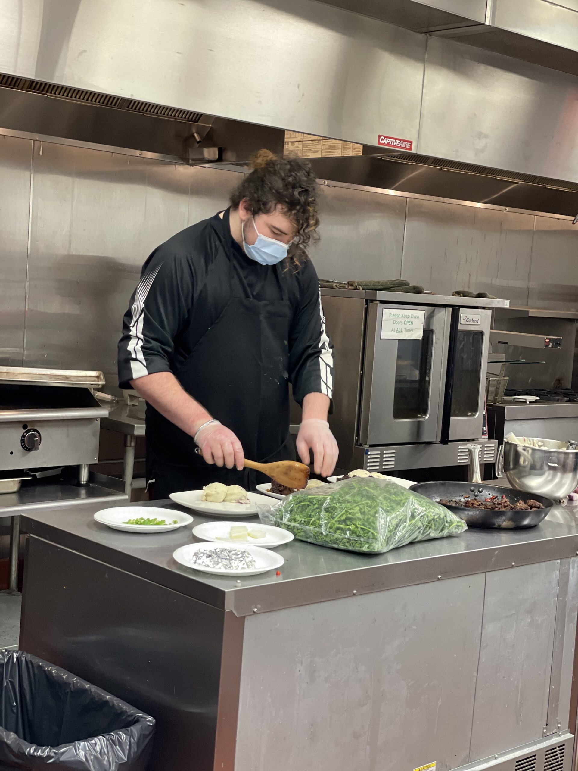 A man in a mask cookingA woman in a red and white jacket cooking in the kitchen. in a busy kitchen.