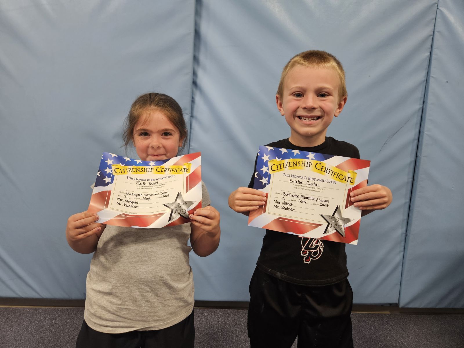 Two children proudly display certificates in front of a wall.