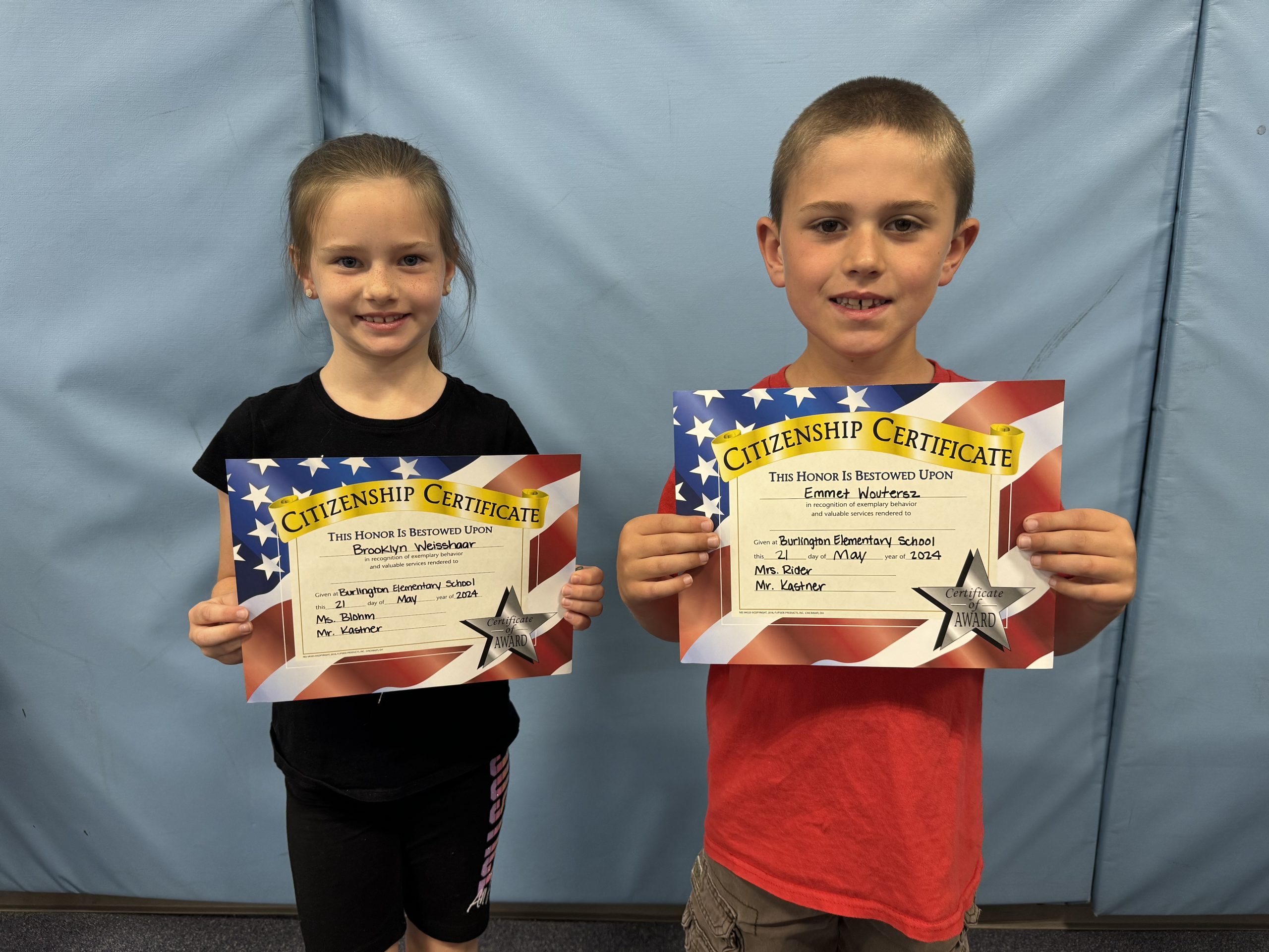 Two children proudly display certificates in front of a wall.