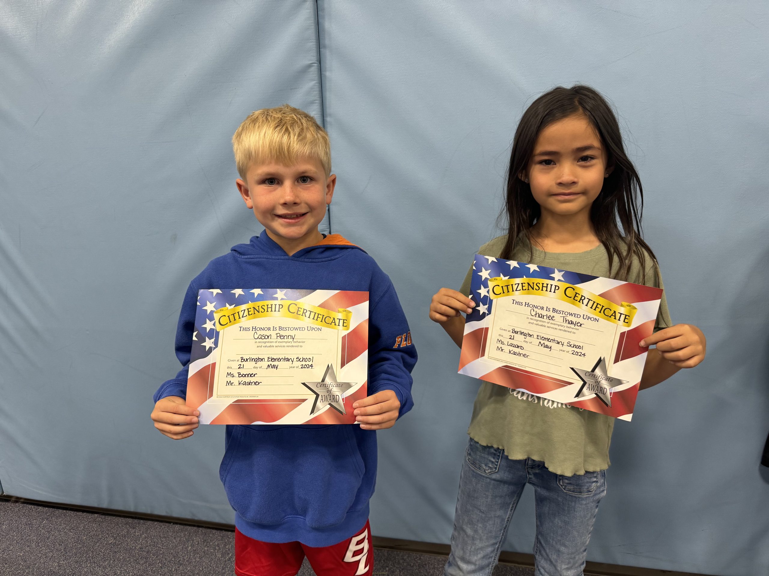 Two children proudly display certificates in front of a wall.