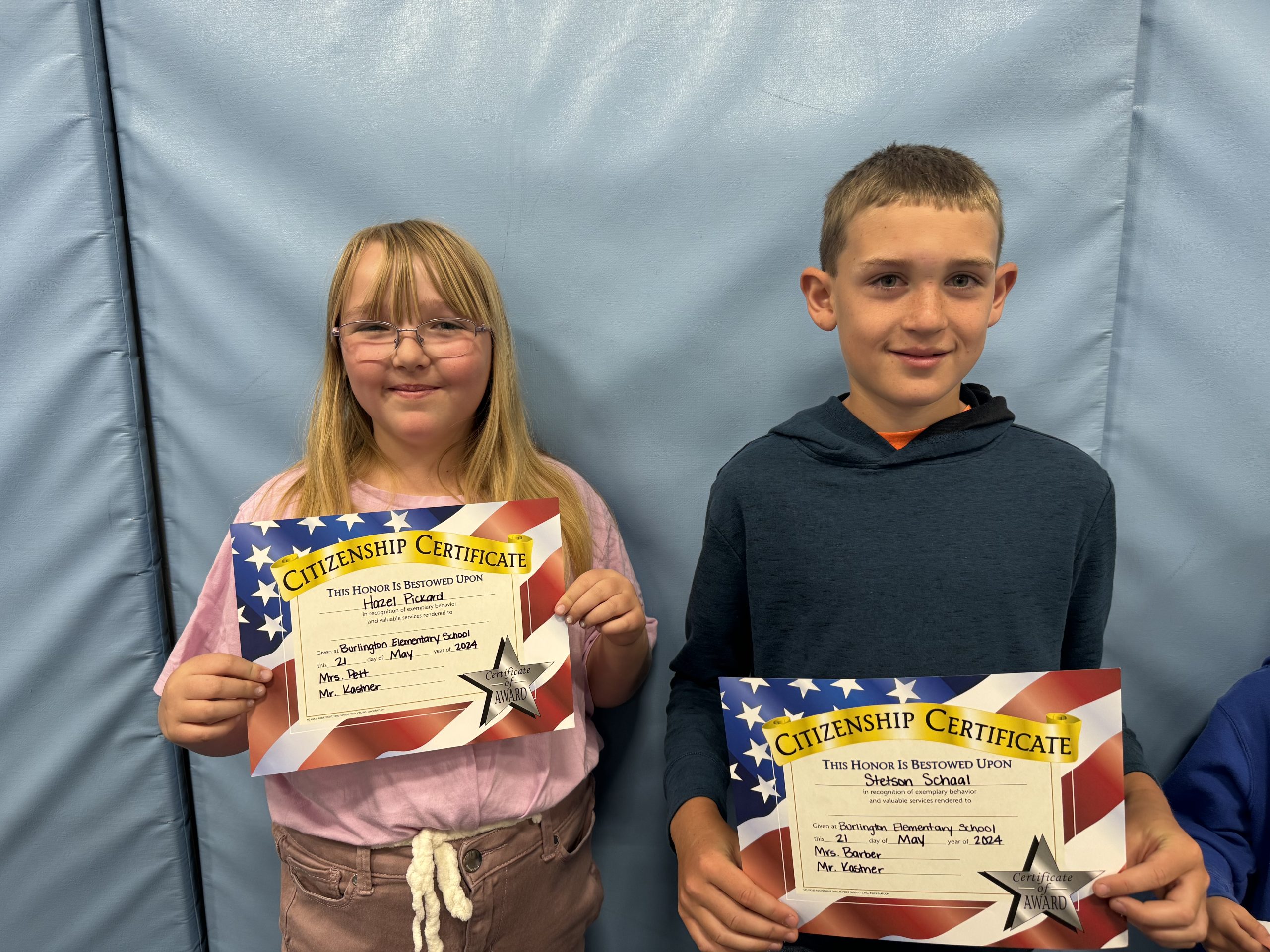 Two children proudly display certificates in front of a wall.
