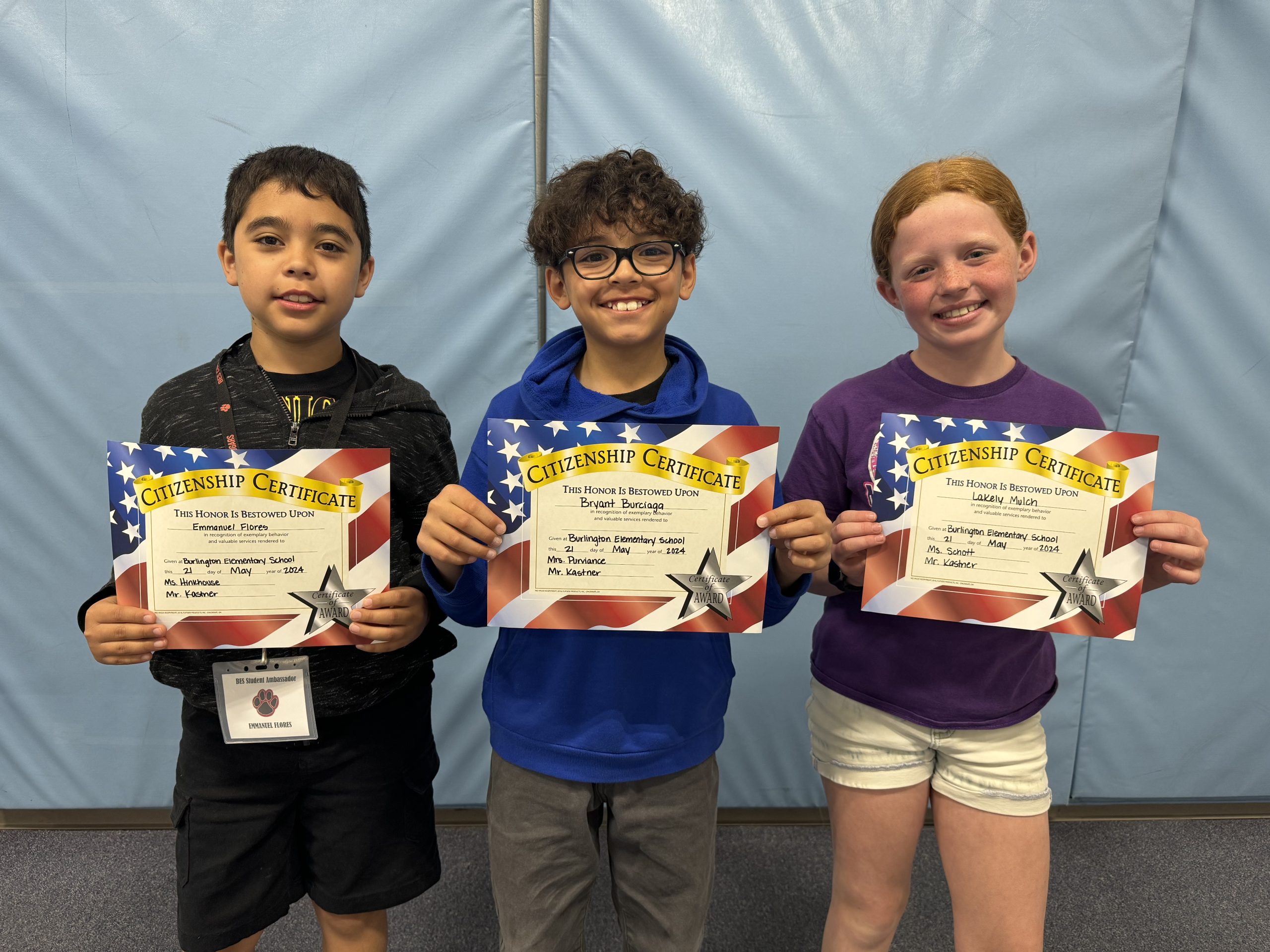 Three children proudly display certificates in front of a wall.