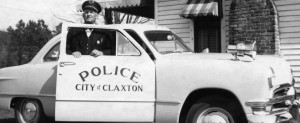 A man in a police uniform stands beside a police car in front of a house, ready to assist the community.