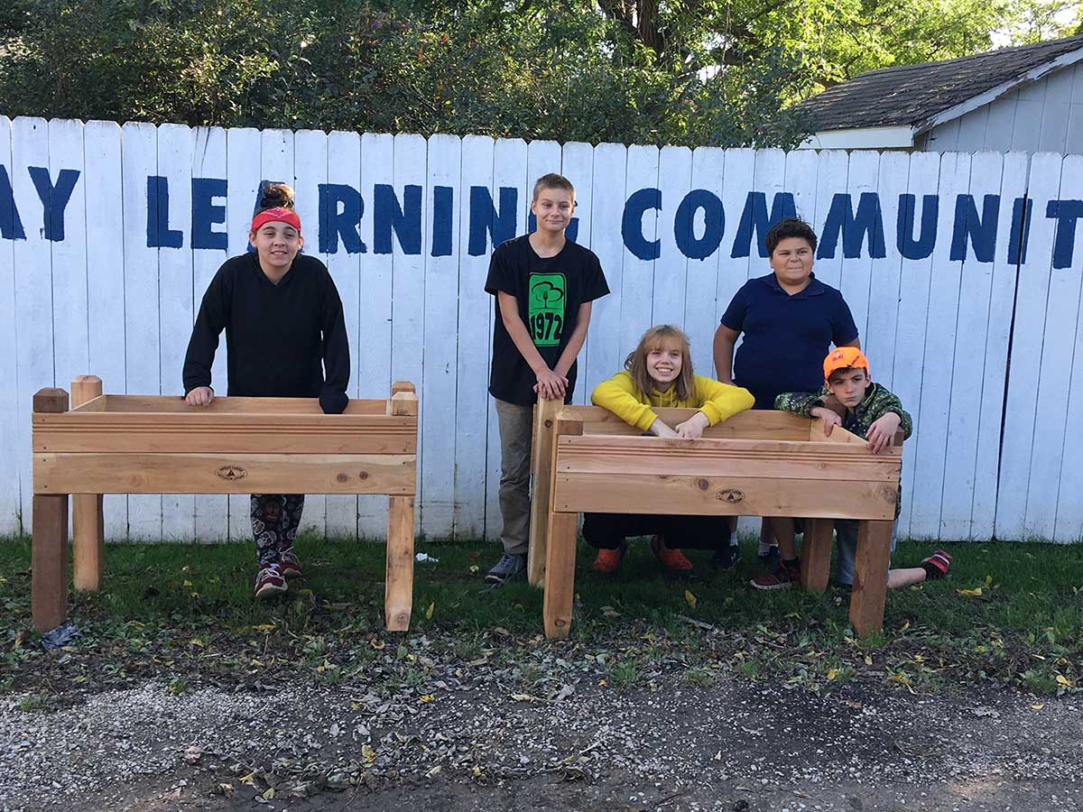 Four people smiling in front of a sign that reads 'Day Learning Community' next to a bench.