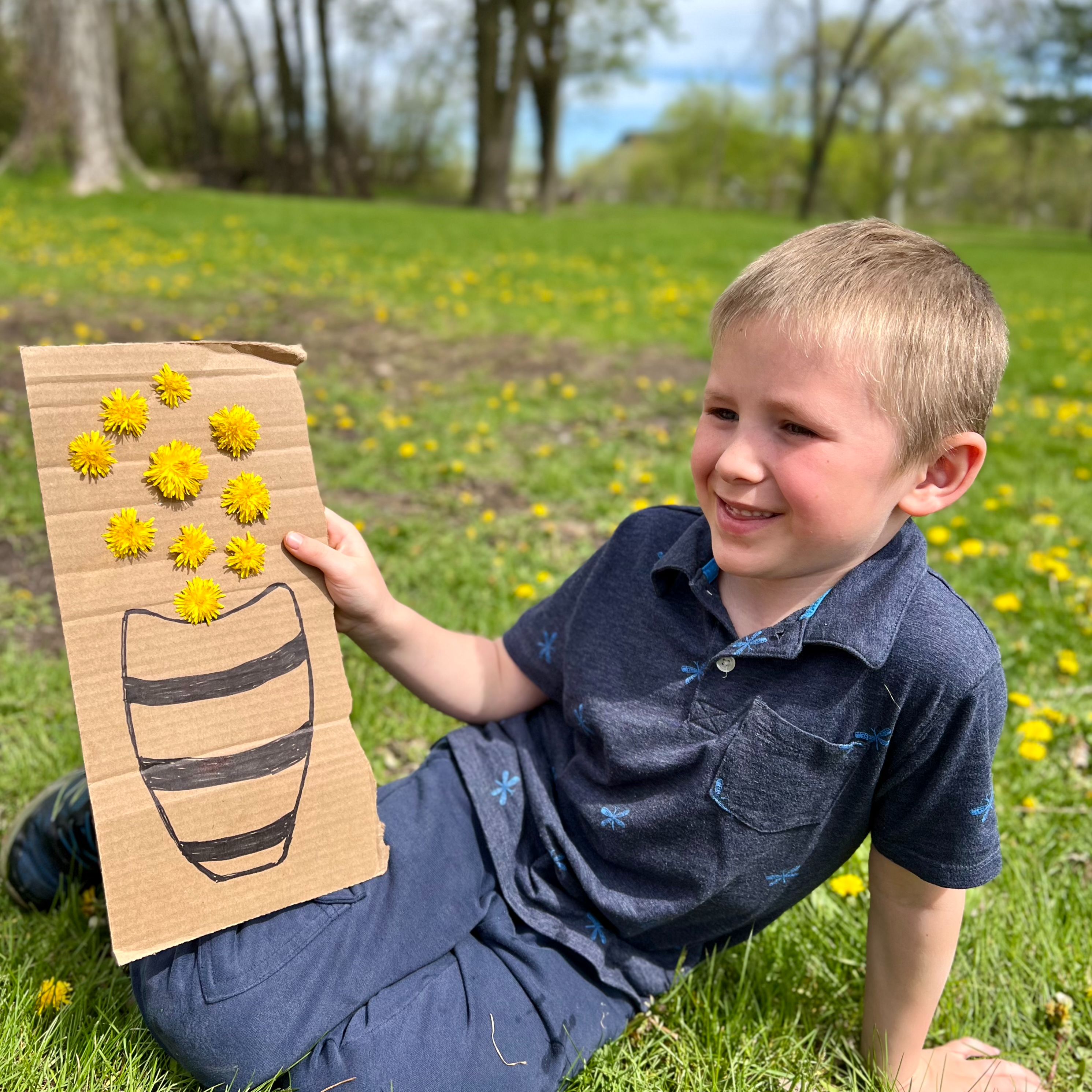 A young boy showing his nature art showing a cardboard with a vace drawn on it with dandelions stuck through the cardboard