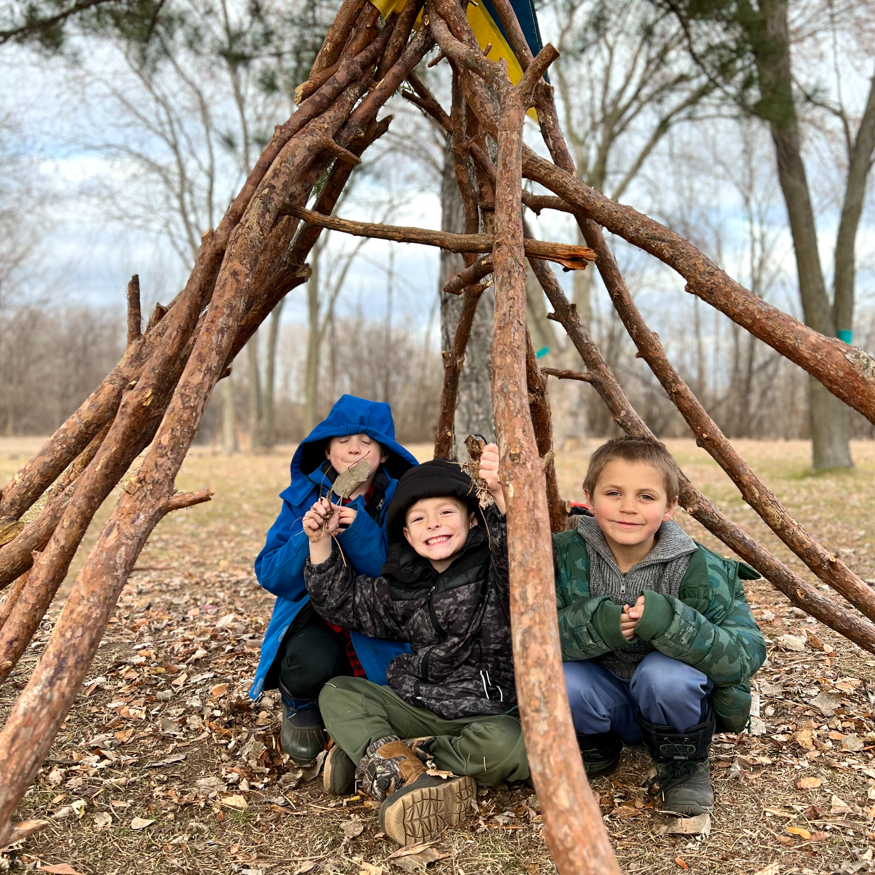 Young boys playing in a teepee made from sticks in the greenspace at Riverway Learning Community