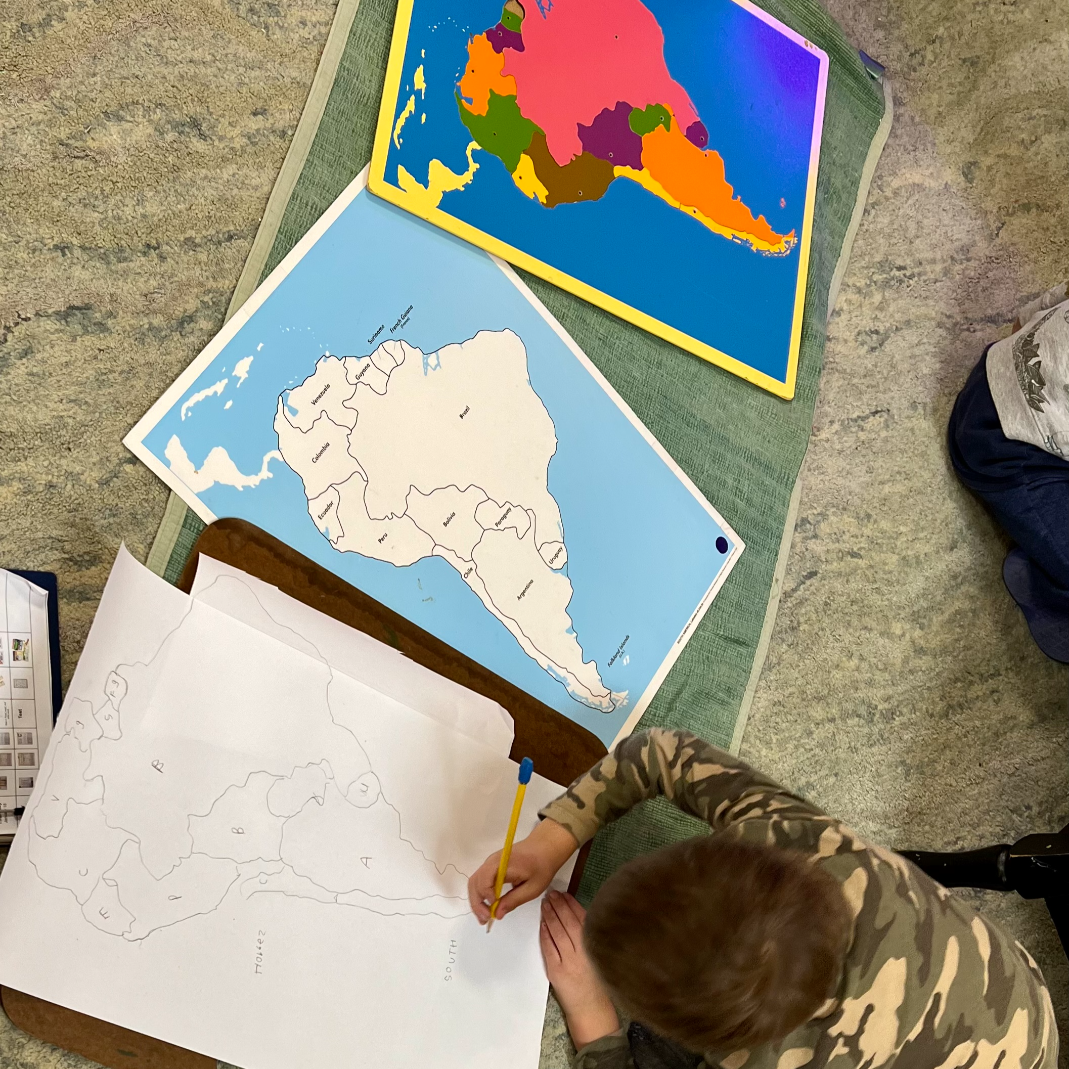 Student working on maps in the Children's House Classroom. Maps. There is a colored map, a blue and white map and the student is working on a pencil lined map drawing.