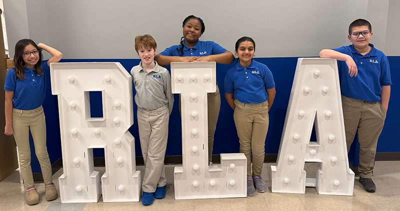 Three elementary school students stand with large letters "R L A"