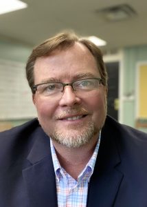 A bearded man wearing glasses sitting at a desk in an office.