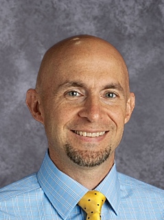 Bald man in blue shirt and yellow tie standing confidently.