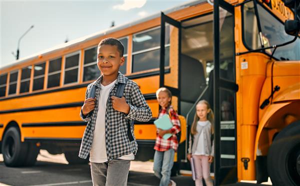 A young boy standing in front of a yellow school bus.