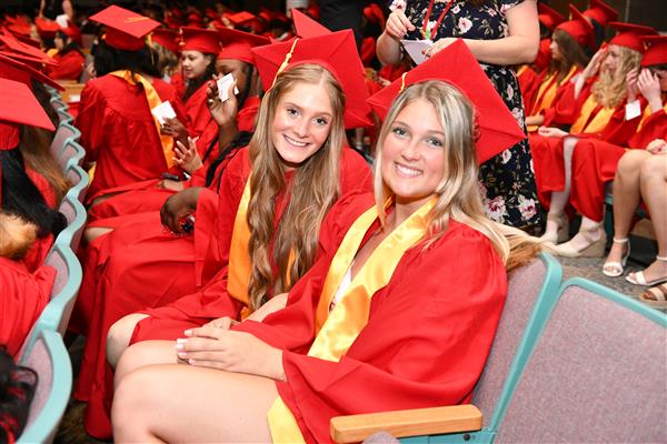 Two girls in red graduation gowns sitting in chairs, smiling and holding diplomas.