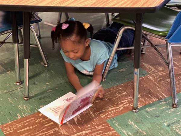 Young girl reading book under table.