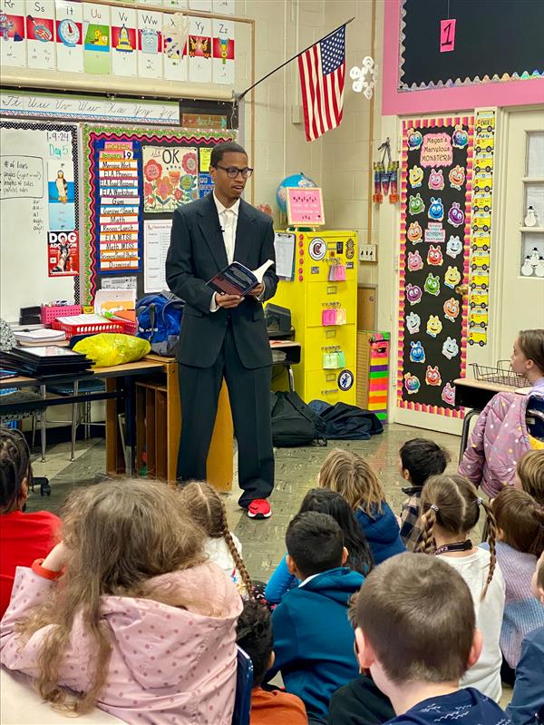 A man in a suit reading a story to a group of children in a classroom.