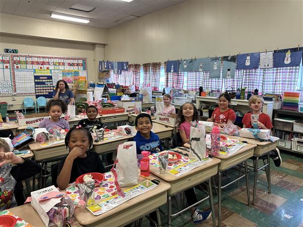 Children sitting at desks in a classroom, listening to the teacher.