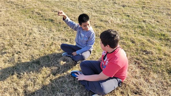 Two boys sitting on ground pointing at something in distance.