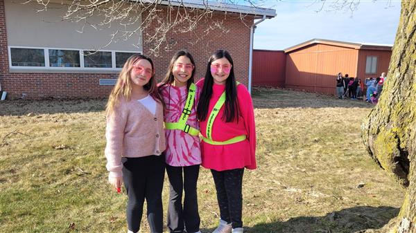 Three girls in pink jackets standing next to a tree.