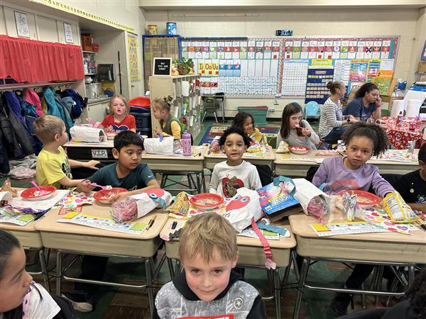 Children sitting at desks in a classroom, listening to the teacher.