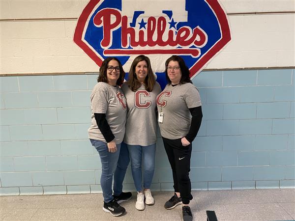 Three women in t-shirts posing in front of a Phillies sign.